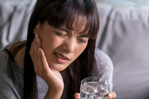 A young Asian woman, clearly in pain, holds her cheek, suggesting she may be experiencing a toothache or sensitive teeth from drinking cold water.
