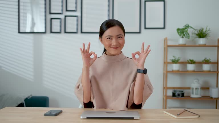 Asian woman happy confident showing OK sign by hand sitting in home office.