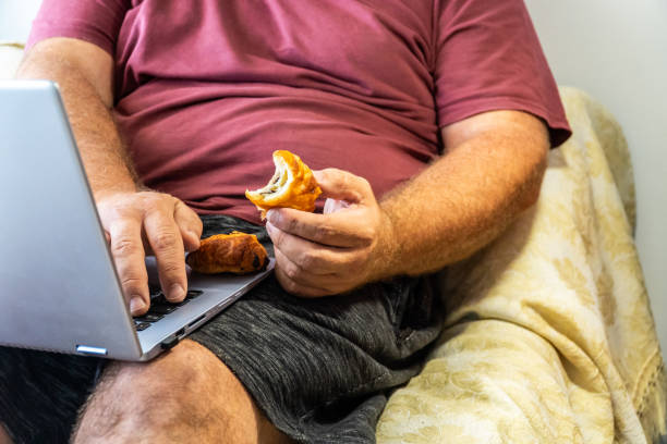overweight obese man eating pastries Unrecognizable person. Sedentary overweight man, sitting eating pastries next to a phone and a laptop excess fat stock pictures, royalty-free photos & images