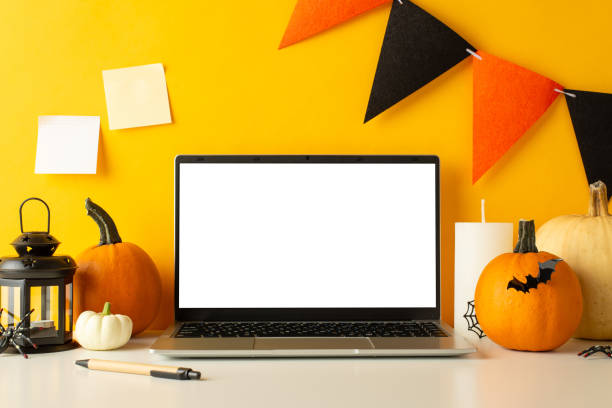istock Ghostly work environment: side view laptop with empty screen amidst spooky decor - pumpkins, bat, candle, spiders, lamp. Yellow wall backdrop with festive flag garland, sticky notes and space for text 1698846229