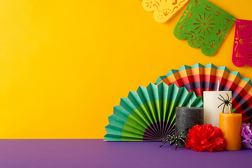 Set mood with side view shot of purple table decked out in Mexican Day of the Dead theme. Vivid fans, flowers, candles, spooky spiders, lively flag garland on yellow wall, space for promo content