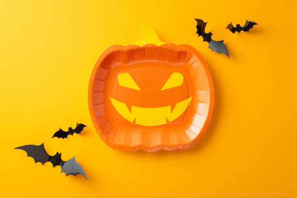 Set the scene for a memorable Halloween event. Top view of themed tableware, jack-o'-lantern plate. Spooky embellishments including bats on orange backdrop