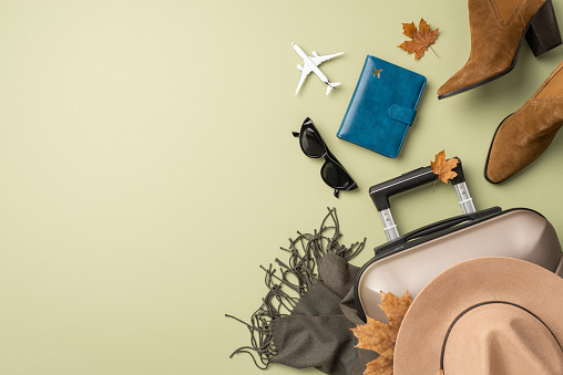 Autumn elegance on a plane journey. Top view of a small aircraft, id passport, fashionable essentials - felt hat, vintage sunglasses, scarf, boots, suitcase, and seasonal decor on light green backdrop