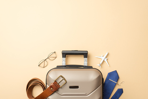 Top-down view of a male's globetrotting essentials: Plane model, necktie with clip, suitcase, belt and glasses, elegantly displayed on a pastel beige surface