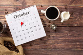 Send your Coffee Day wishes. Top view of calendar with highlighted 1st October, bag with roasted beans, espresso, milk jar, coffee pot, barista's scoop on wooden desk backdrop, ready for message or ad