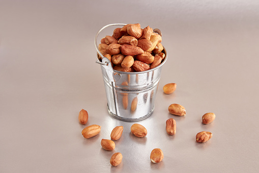 A small metal bucket filled with raw peanuts on a colored background.