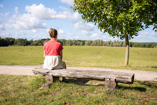 An elder woman sitting on a bench during the day in summer in the nature near a meadow looking to the distance.