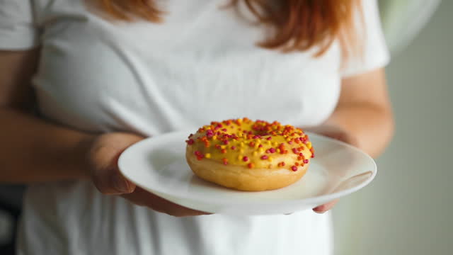 Woman wearing white shirt holding and showing a pale plate of sugar glazed donut in a modern cafe. Enjoyment female lifestyle. Focus on hand and donut.