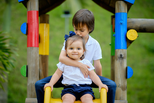 Two happy siblings, brother hugging his sister and kissing her on the head. They are in a wooden playground in a free and natural space