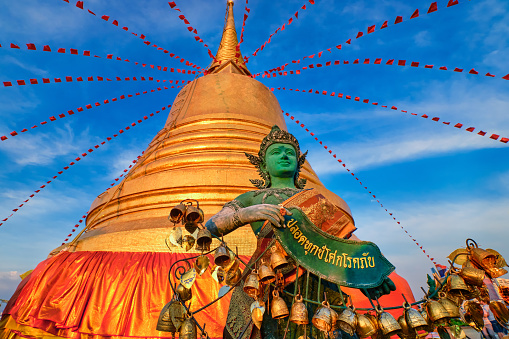Statue of green-skinned Thai angel holding roll with message in Thai stand in front of big golden stupa in sunlight and at blue sky with clouds in Buddhist temple. Decorations with bells, golden leaves and red flags. Translation: free from sorrow