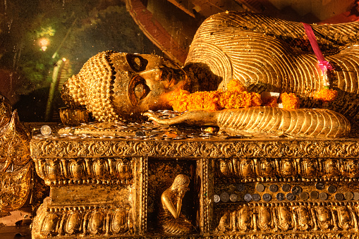 Statue of reclining Buddha covered with golden leaves, coins and garlands of flowers as offerings in soft sunlight. Traditional image, rituals of prayer, worship and blessing. Buddhism, religion, Theravada, Thailand, sacred.