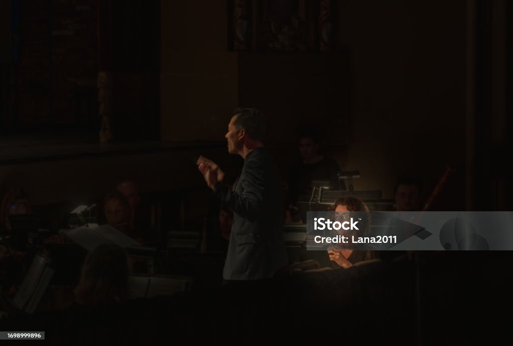 View or orchestra oboe player with a silhouette of orchestra conductor in foreground and vague figures of musicians in background during concert in dark hall Musical Instrument Stock Photo