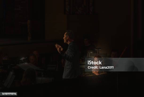 View Or Orchestra Oboe Player With A Silhouette Of Orchestra Conductor In Foreground And Vague Figures Of Musicians In Background During Concert In Dark Hall Stock Photo - Download Image Now