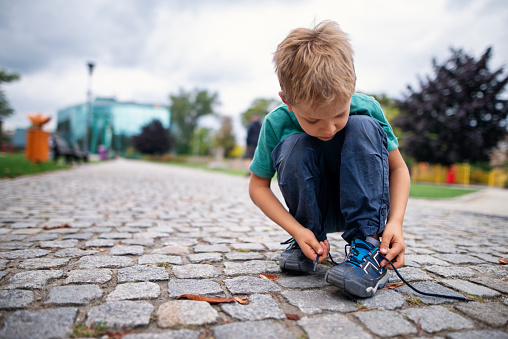 Little boy is tying a shoelace on the paved walkway.