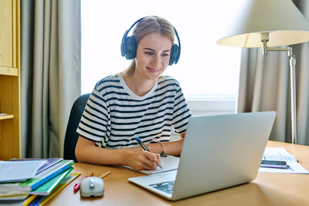 istock Girl teen student in headphones sitting at desk using computer writing in study notebook 1699081678