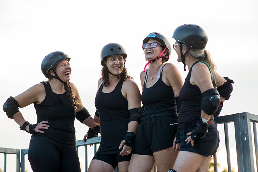 Four friends, three female and one genderqueer are all wearing black clothing, and protective helmets, elbow pads, kneepads and wrist guards as they pause at the top of a ramp at a skateboard park while practicing roller skating together for roller derby.