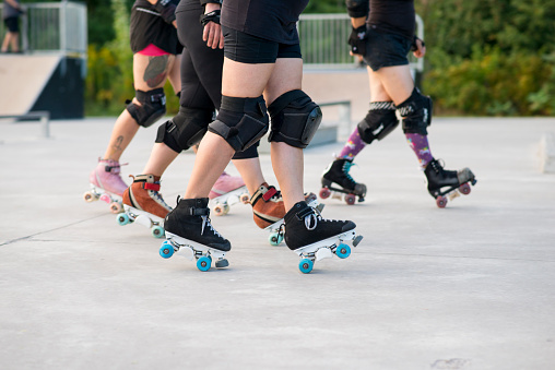 Four roller skating friends are lined up in sync doing the same manual trick angling both skates as they ride forward. They are all wearing black clothing and kneepads for protection. They are practicing at the skateboard park outdoors in the summer.