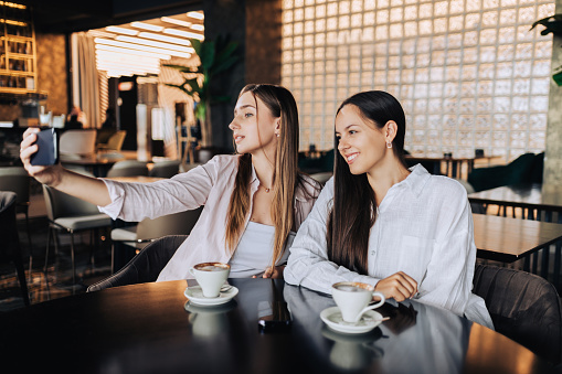 Two happy girls are sitting in a cafeteria and taking self-portraits during their coffee time. Two girls are capturing selfies while sitting in a cafe during their coffee break.