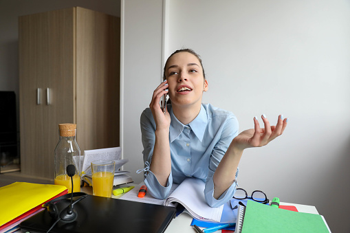 A young businesswoman with a phone in her hand in a blue shirt is sitting on a chair in the office and having a conversation. in the background a white wall