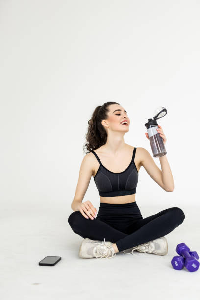 istock Dehydration After Workout. Young woman with white towel on neck drinking mineral water from bottle, taking break training sitting on floor on white background 1699111734