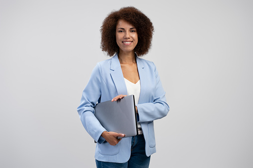 Young African American woman with afro hairstyle wearing smart formal wear standing isolated on grey , carrying laptop computer, smiling, female office employee. Successful business concept
