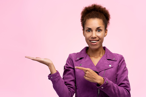 African American charming woman in purple jacket pointing with forefinger at imaginary copy space on her hand palm up, smiling a cheerful toothy smile looking at camera, isolated over pink background