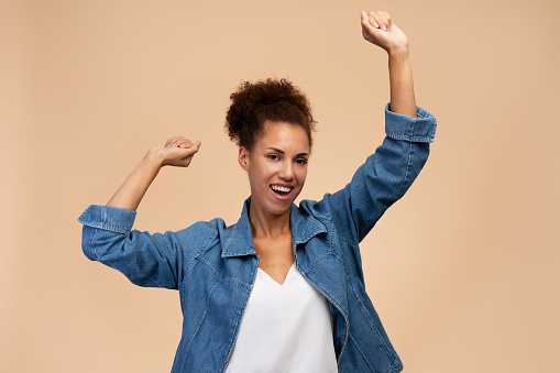 Pretty multi-ethnic woman with afro hairstyle, wearing blue casual denim, freely moving, dancing and smiling a cheerful smile looking at camera, isolated beige background. People Leisure Lifestyle