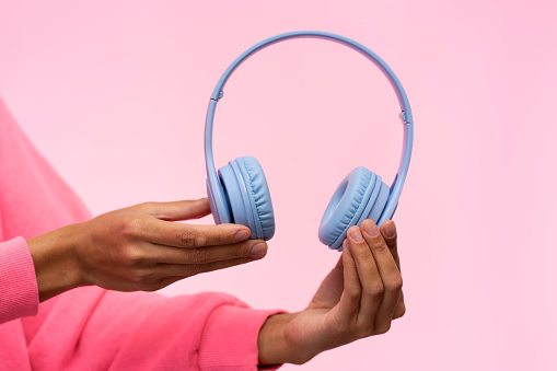 Close-up of female hands holding modern wireless blue headphones, isolated over pink background. Copy advertising space. Audio headset.