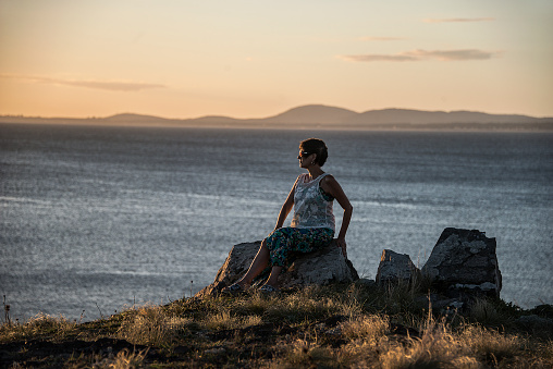 Mature woman sitting on a rock in Punta Ballena, at sunset, looking towards the sea and with the ocean and the hills of Piriápolis behind, Uruguay.