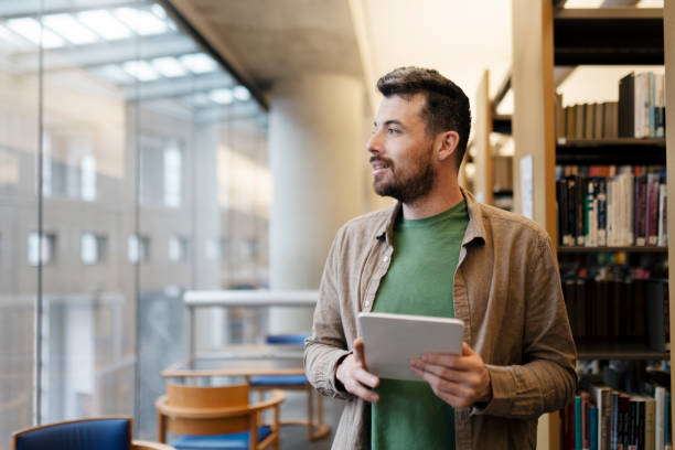 Handsome latin man holding digital tablet looking away. Middle aged student studying in library Portrait of pensive handsome latin man holding digital tablet looking away, copy space. Middle aged student studying in library, education concept mature library stock pictures, royalty-free photos & images