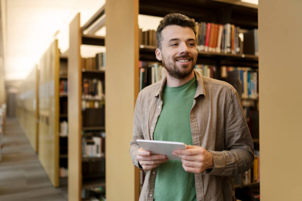 istock Smiling, handsome hispanic man standing near bookshelf, holding digital tablet, reading e books 1699272551