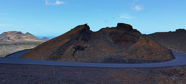 This captivating image centers on a black road winding around a brown-black volcanic crater. In the distance, a cluster of volcanoes frames the view. The ocean and an endless blue sky blend harmoniously into a single hue on the horizon.