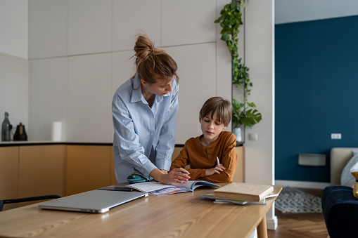 Young mother helping child son with homework at home, sitting together at kitchen table. Happy schoolboy learning foreign language with private teacher tutor at home. Homeschooling concept