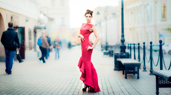 Young brunette woman wearing remarkable red dress is standing on city streets and looking into camera.