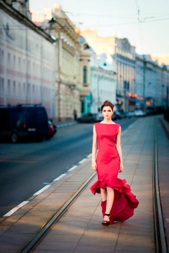 Young brunette woman wearing remarkable red dress is calmly walking on city streets and looking into camera.