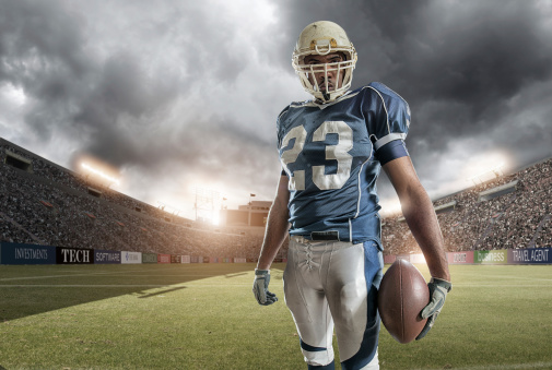 american football player standing holding ball in full floodlit stadium under stormy sky at sunset