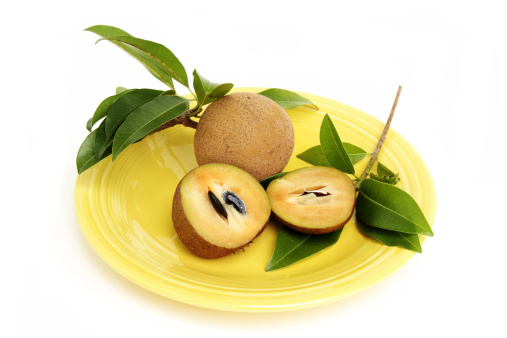 Sapodilla, Manikara Zapota,  is a popular fruit in Mexico, South America, the Caribbean, and Middle Eastern countries like India and Pakistan. Also known as sapota, zapota, and chikoo. This image shows a sliced fuit in foreground with seeds in one side. A  whole, uncut fruit is in the background, The fruit is on a yellow platter and surrounded by several sapodilla tree leaves. Fruit is very sweet when ripe and has a malty, caramel flavor.
