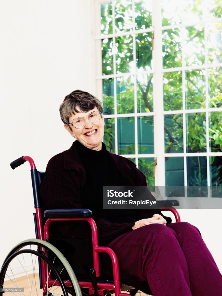 Smiling senior woman sitting in wheelchair A elderly woman sits in a wheelchair smiling. She seems content with her lot or perhaps she's recovering her health. Through a window behind her is a spring-green garden. 60-69 Years Stock Photo
