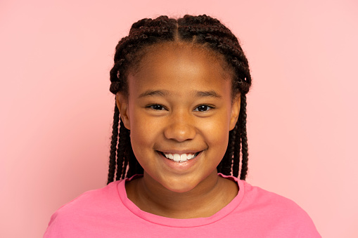 Portrait of cute smiling African American girl wearing stylish hairstyle, dreadlocks, pigtails wearing casual pink t shirt standing isolated on pink background, looking at camera, closeup