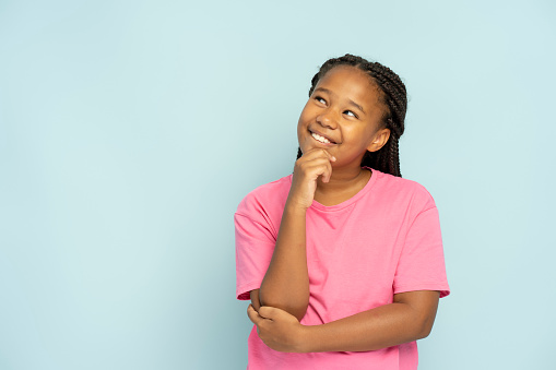 Beautiful smiling Nigerian girl with stylish hairstyle in casual clothes standing looking away, thinking isolated on blue background. Positive child posing. Childhood concept
