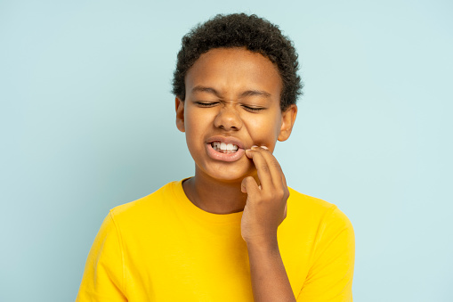 Upset handsome African American boy with closed eyes touching cheek, having toothache wearing stylish yellow casual t shirt isolated on blue background. Dental treatment concept