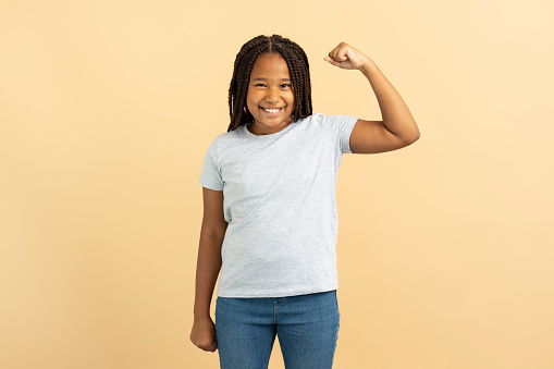 Portrait of smiling positive Nigerian girl with stylish hairstyle, dreadlocks, in casual clothes showing muscles isolated on beige background. Healthy lifestyle concept