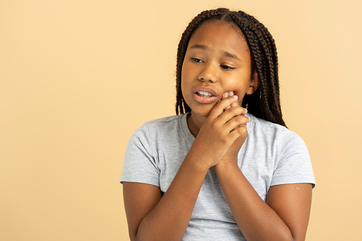 Upset beautiful African American girl with stylish pigtails touching cheek, toothache, looking away isolated on beige background. Sad female child. Dental care concept