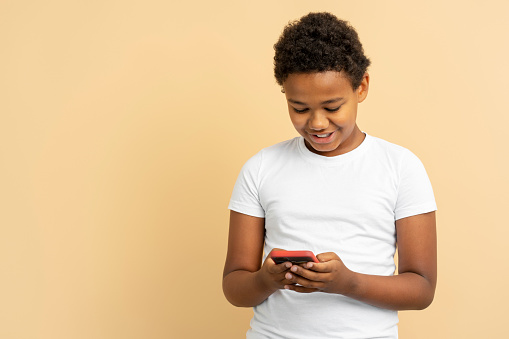 Portrait of smiling handsome African American boy in white t shirt holding mobile phone, text message, ordering, online shopping isolated on beige background, copy space. Social media concept