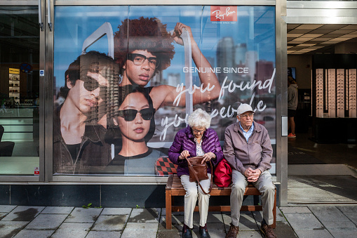 Stockholm, Sweden Aug 15, 2023 A senior couple sitting on a bench are juxtasposed with a sunglass advertisement in a shop window of young people on Norrlandsgatan.