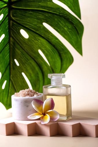 SPA composition, essential oil , salt scrub and frangipani flower on the podium on beige background with palm leave . Beauty and wellness centre concept .