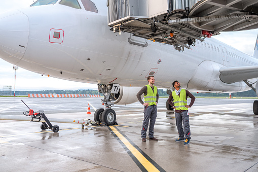 Multiracial male coworkers standing outdoors next to a white plane. They are wearing reflective vests and looking at a jet bridge.