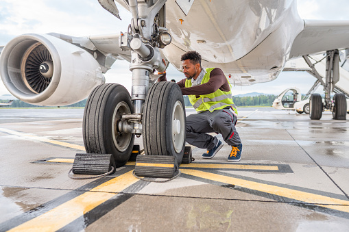 Handsome mid adult Hispanic male employee working under an aircraft. He is checking the wheels and wearing a reflective vest.