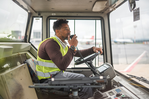 Side view of a mixed race male worker driving a truck at the airport. He is talking with a walkie talkie. He is wearing a reflective vest.