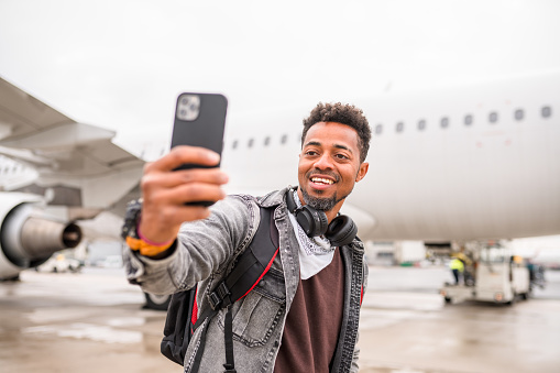 Cheerful Black male passenger taking a selfie at the airport. Carrying a backpack and headphones.